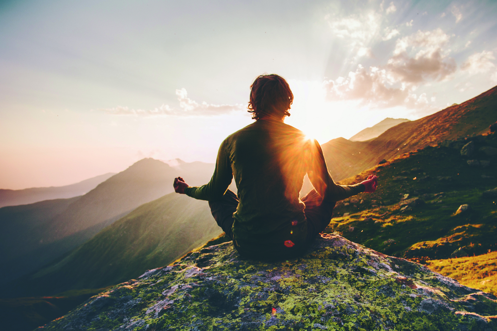 Ein Mann sitzt in Yoga-Stellung auf einem Felsen und schaut mit gehobenen Armen in die Morgensonne